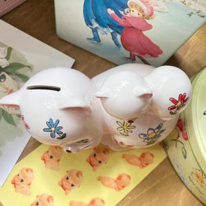 May include: White ceramic piggy bank with a floral design. The piggy bank has a slot on the top for coins and is decorated with colourful flowers.