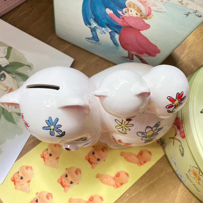 May include: White ceramic piggy bank with a floral design. The piggy bank has a slot on the top for coins and is decorated with colourful flowers.