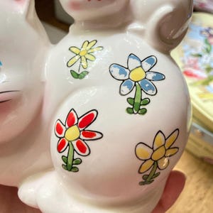 May include: White ceramic piggy bank shaped like a cat with colourful flower designs. The flowers are red, yellow, blue, and green.