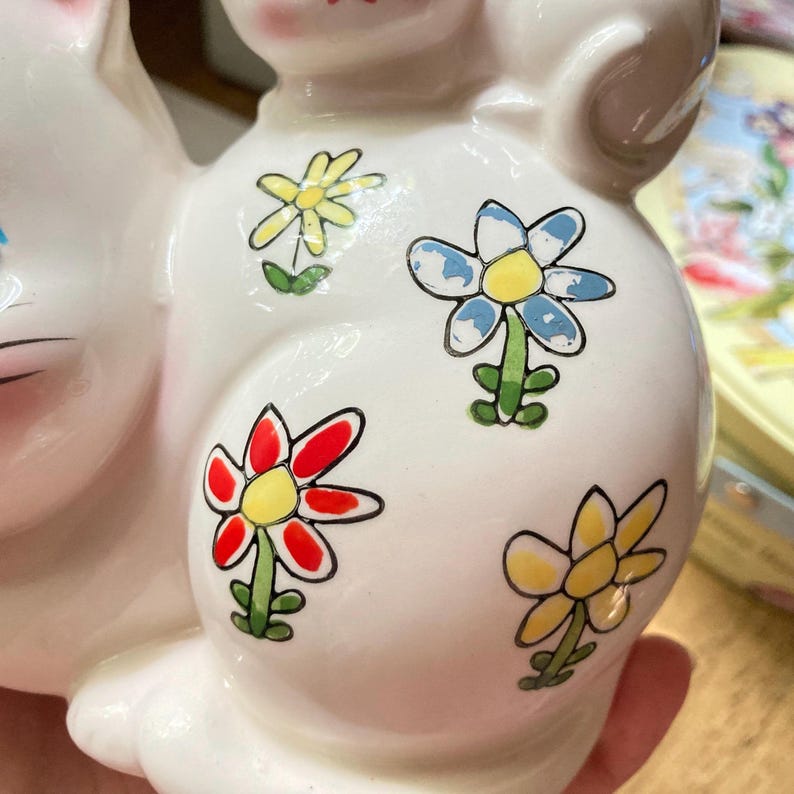 May include: White ceramic piggy bank shaped like a cat with colourful flower designs. The flowers are red, yellow, blue, and green.