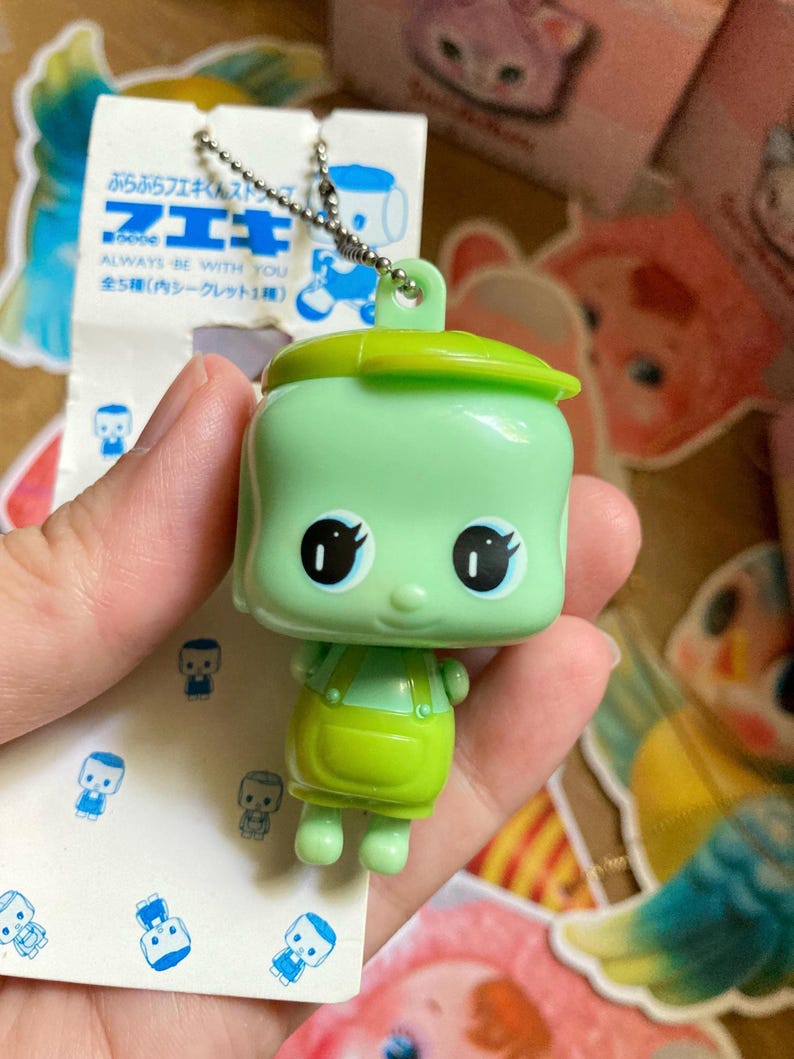 May include: A green, square-shaped keychain figure with large black eyes and a green lid. The figure is wearing green overalls. The attached tag reads "ALWAYS BE WITH YOU" and includes Japanese text.