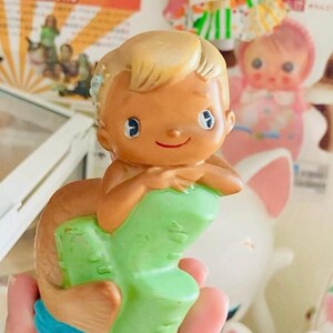 May include: A vintage mermaid figurine with a tan complexion, blonde hair, and blue eyes. The mermaid is perched on a green rock formation with a brown tail and blue water base. The figurine is held in a hand, with other toys in the background.