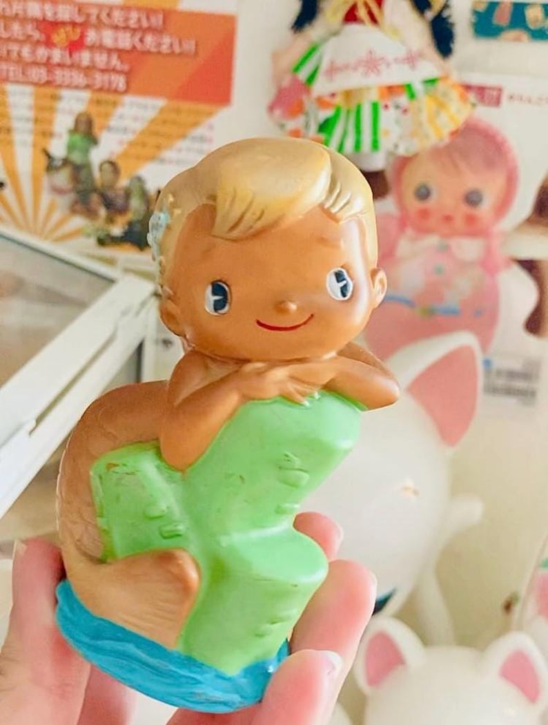 May include: A vintage mermaid figurine with a tan complexion, blonde hair, and blue eyes. The mermaid is perched on a green rock formation with a brown tail and blue water base. The figurine is held in a hand, with other toys in the background.