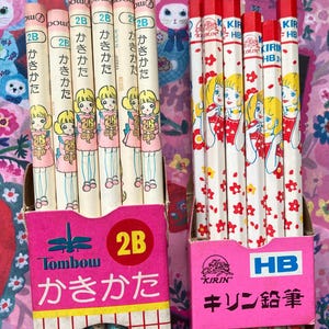Puede incluir: Dos cajas de l&aacute;pices japoneses vintage. Una caja contiene l&aacute;pices con una ni&ntilde;a sosteniendo un oso de peluche, la otra con una ni&ntilde;a con un vestido rojo. Las cajas son rosas con texto japon&eacute;s y el grado del l&aacute;piz es 2B y HB.