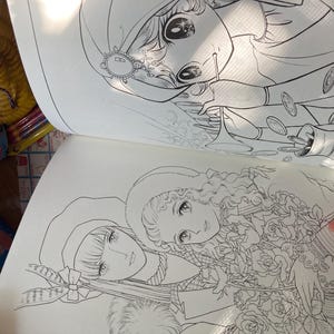 May include: Open coloring book pages featuring detailed line art illustrations of manga-style characters. One page shows a woman with coins, the other shows two women with flowers. The pages are open, revealing the intricate drawings.