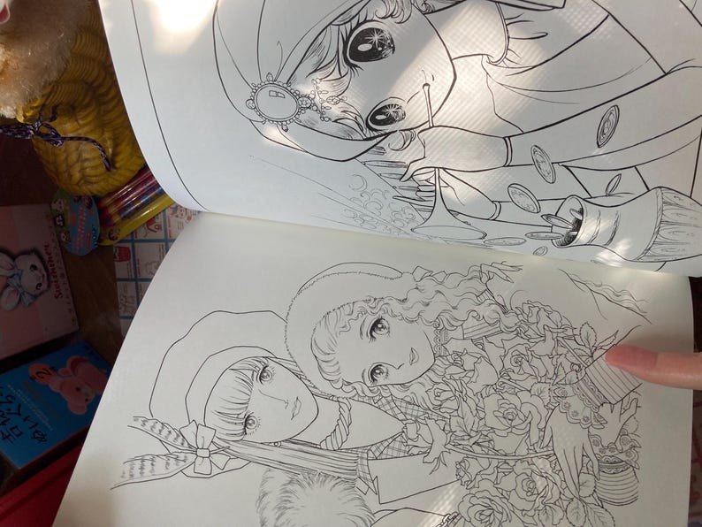 May include: Open coloring book pages featuring detailed line art illustrations of manga-style characters. One page shows a woman with coins, the other shows two women with flowers. The pages are open, revealing the intricate drawings.
