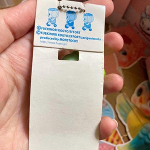May include: A white product tag with three blue cartoon figures and text. The tag is attached to a small green object. The text includes "FUEKINORI KOGYO/EFFORT" and "produced by MONSTOCK!!".