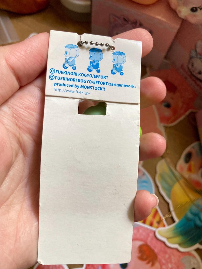 May include: A white product tag with three blue cartoon figures and text. The tag is attached to a small green object. The text includes "FUEKINORI KOGYO/EFFORT" and "produced by MONSTOCK!!".