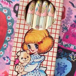 Puede incluir: Juego de l&aacute;pices vintage con una ni&ntilde;a de dibujos animados con un cachorro, con un vestido azul con lunares. La caja de l&aacute;pices tiene un patr&oacute;n de cuadr&iacute;cula roja y blanca. Los l&aacute;pices tienen un dise&ntilde;o de mu&ntilde;eca.