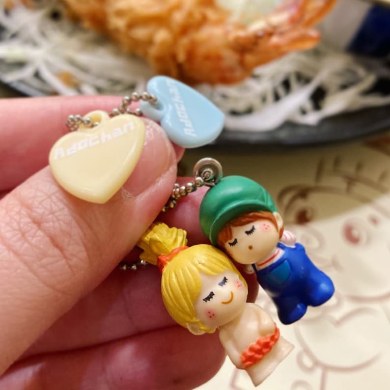 May include: A collection of miniature keychains featuring a heart-shaped charm with the text "Adochan", and two small figurines of a boy and a girl. The boy wears a green hat and blue overalls. The girl has blonde hair.