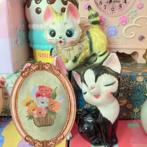 May include: A ceramic cat figurine with black and white fur, closed eyes, and a pink nose, positioned next to an oval-shaped frame featuring a painted floral arrangement in a basket, with a yellow and grey cat figurine visible in the background.