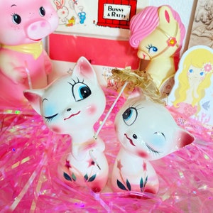 May include: Two white ceramic kittens with pink accents and winking faces. One kitten holds a small, sheer, white umbrella with a gold handle. The kittens are sitting on a pink and white fluffy surface.