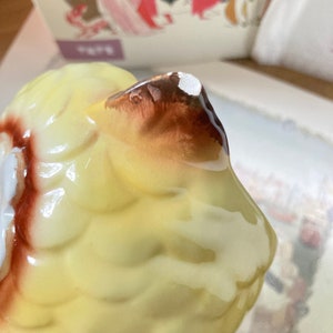 May include: Close-up of a ceramic owl figurine. The owl has yellow feathers with brown and white accents. The figurine has a glossy finish and is partially visible against a blurred background.
