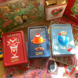 Könnte beinhalten: Eine Sammlung dekorativer Metallboxen im Vintage-Stil. Die Dosen zeigen Illustrationen von Desserts wie Parfaits, Puddings und einem blauen Soda. Andere Dosen zeigen Gartenszenen und ein Mädchen mit einem Herz.