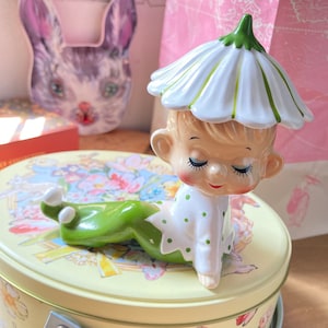 May include: A ceramic figurine of a child with a daisy-shaped hat and green pants. The child is lying down with closed eyes and a serene expression. The figurine is on a floral-patterned tin.