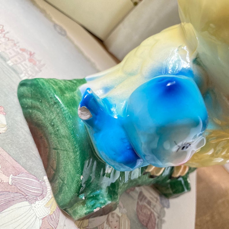 May include: A vintage ceramic figurine of a bluebird perched on a green branch. The bird has a blue head and yellow body. The figurine has some visible wear and tear, including a chip on the bird's wing.