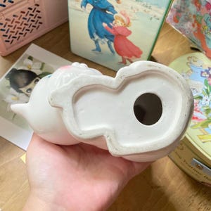 May include: White ceramic figurine of a cat with a hole in the bottom. The cat is sitting up with its head tilted to the side. The figurine is approximately 15 cm tall.