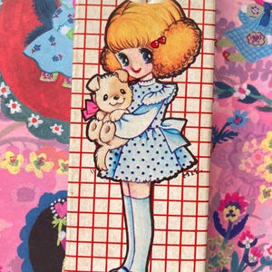 Puede incluir: Una caja de l&aacute;pices vintage con una ni&ntilde;a de dibujos animados con un vestido azul de lunares y un osito de peluche. La caja tiene un patr&oacute;n de cuadr&iacute;cula roja. Los l&aacute;pices son visibles en la parte superior. El tema es retro y nost&aacute;lgico.