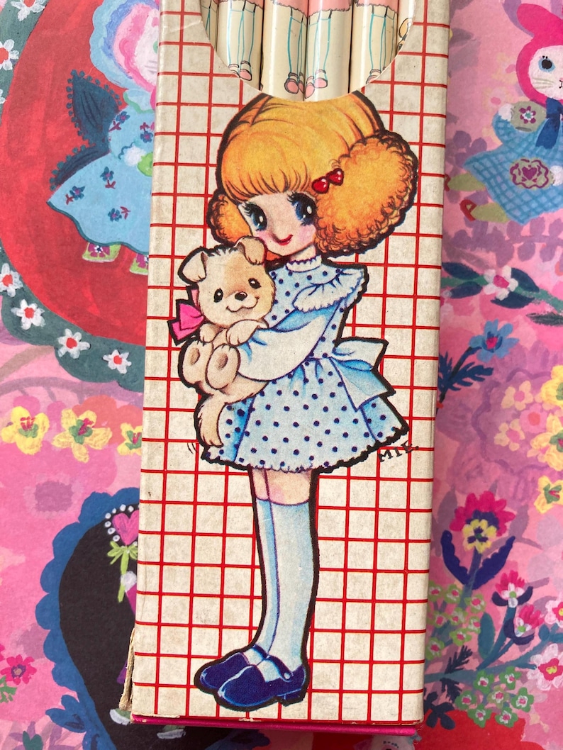 Puede incluir: Una caja de l&aacute;pices vintage con una ni&ntilde;a de dibujos animados con un vestido azul de lunares y un osito de peluche. La caja tiene un patr&oacute;n de cuadr&iacute;cula roja. Los l&aacute;pices son visibles en la parte superior. El tema es retro y nost&aacute;lgico.