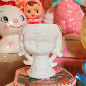Peut inclure: Figurine en céramique blanche représentant une jeune fille aux yeux fermés et aux couettes. La figurine a une base évasée et des détails floraux. Elle est posée sur une boîte décorative.