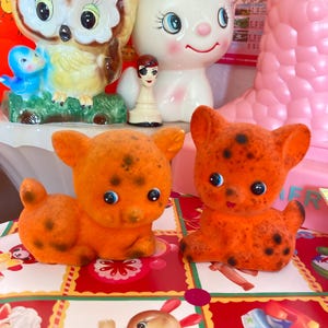 Op de afbeelding: Twee oranje, bevlokte kattenbeeldjes met zwarte vlekken en blauwe ogen. De beeldjes staan op een kleurrijk patroon. Andere keramische beeldjes, waaronder een uil en een witte kat, zijn zichtbaar op de achtergrond.