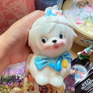 Puede incluir: Figura de perro de cerámica blanca con un lazo azul y un sombrero blanco. El perro tiene la nariz rosa y los ojos azules.