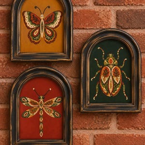 May include: Three arched wall plaques featuring painted insects. One shows a butterfly on a mustard background, one a dragonfly on a red background, and one a beetle on a dark green background. Each piece is encased in a distressed black frame.