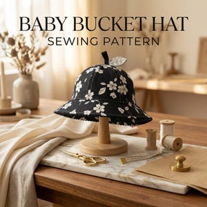 May include: A black bucket hat with a white floral pattern and a small fabric leaf detail on top. The text BABY BUCKET HAT SEWING PATTERN is displayed above the hat. The hat is positioned on a wooden stand with sewing tools nearby.