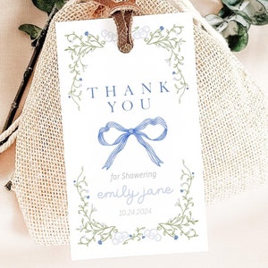 Puede incluir: Una etiqueta de agradecimiento blanca con un borde floral azul y un lazo azul. La etiqueta dice "Gracias por duchar a Emily Jane 10.24.2024".