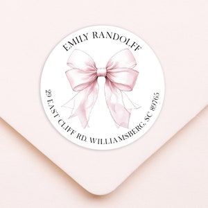 May include: A round sticker with a watercolor illustration of a pink bow. The sticker has the name "EMILY RANDOLFF" at the top and address details below. The sticker is on a light pink envelope.
