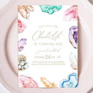 May include: A white invitation card with watercolor gemstone illustrations in shades of pink, blue, green, and gold. The text reads "Our Little Gem Charlotte is Turning One" with event details.