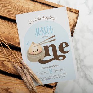 May include: A blue and white invitation card with a cartoon dumpling illustration. The text reads "Our little dumpling Joseph is turning one. Join us to celebrate September 23, 3pm-6pm, 18 Dumpling way, Kenilworth AZ 08765, rsvp by 8/15 to Sam at 897-456.1234"