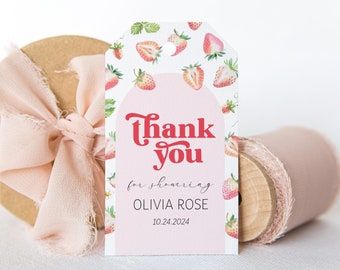 May include: A rectangular paper gift tag featuring a strawberry pattern and a pink arch design. The text reads Thank you for showering OLIVIA ROSE 10.24.2024 in red and black ink. The tag has a hole at the top and is positioned in front of a wooden spool.