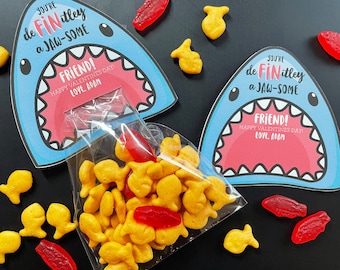May include: Editable printable Valentine's Day card featuring a blue shark with an open mouth and the text "You're de Fin itley a Jaw-Some Friend! Happy Valentine's Day! Love, Adam." The card is designed to be used with a bag of goldfish crackers.