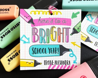 May include: Editable printable gift tag with a colorful "Here's to a Bright School Year!" message. The tag is designed to look like a piece of notebook paper with a grid pattern. The text "From: Alexandra" is included at the bottom of the tag.