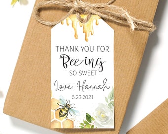 May include: A rectangular white gift tag with a twine string featuring a yellow honey drip design at the top. The text reads THANK YOU FOR Bee-ing SO SWEET Love, Hannah 6.23.2021. It includes a watercolor illustration of a bee, honeycomb, and white flowers.
