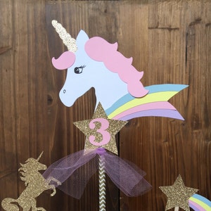 May include: Unicorn cupcake toppers with a rainbow, star, and the number 3. The toppers are on cupcakes with white frosting and are surrounded by confetti.