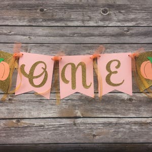 K&ouml;nnte beinhalten: Rosa-goldene Girlande mit dem Wort "eins" in goldenen Glitzerbuchstaben und K&uuml;rbis-Ausschnitten. Die Girlande ist mit goldenen Band befestigt.