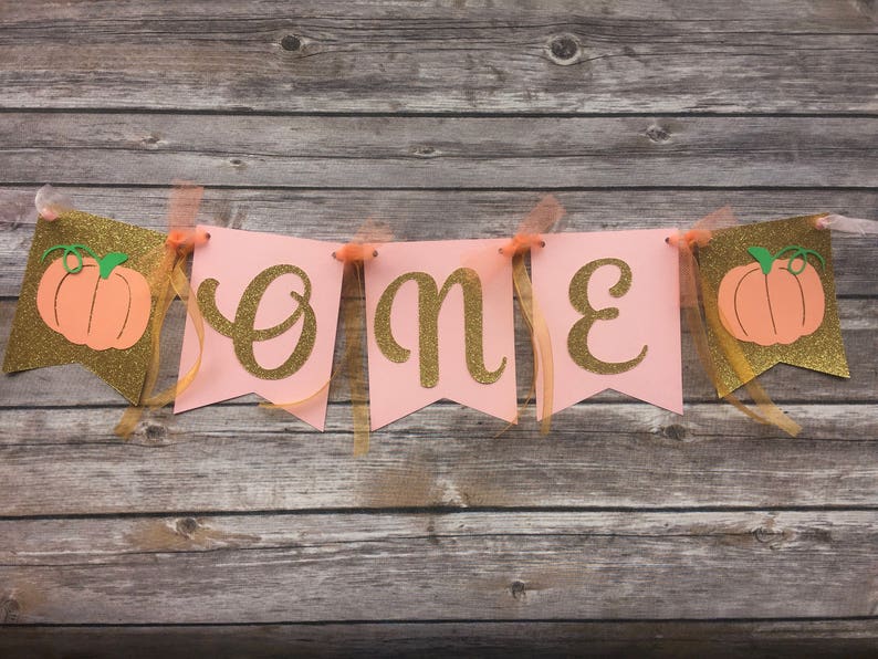 K&ouml;nnte beinhalten: Rosa-goldene Girlande mit dem Wort "eins" in goldenen Glitzerbuchstaben und K&uuml;rbis-Ausschnitten. Die Girlande ist mit goldenen Band befestigt.