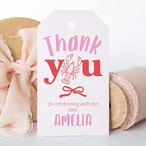 May include: A white gift tag with pink and red text that says "Thank You" with a lobster illustration. The tag also includes the text "for celebrating with me! love, AMELIA" and a small bow illustration. A roll of pink ribbon and a wrapped gift are in the background.
