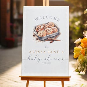 Może przedstawiać: Witamy znak na baby shower na sztaludze. Znak zawiera akwarelową ilustrację bułek cynamonowych w naczyniu do pieczenia, z tekstem "WELCOME to ALYSSA JANE'S baby shower 11.23.2025."