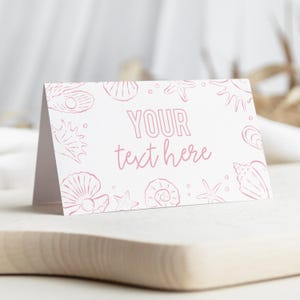May include: A folded white card with pink seashell illustrations and the text "YOUR text here." The card is on a wooden surface. The design includes various seashell and starfish outlines.