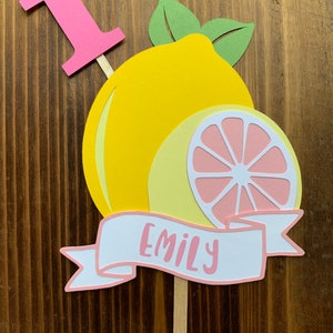 May include: A pink number one cake topper with a yellow lemon and a pink lemon slice. The topper also has a white banner with the name "Emily" written in pink.