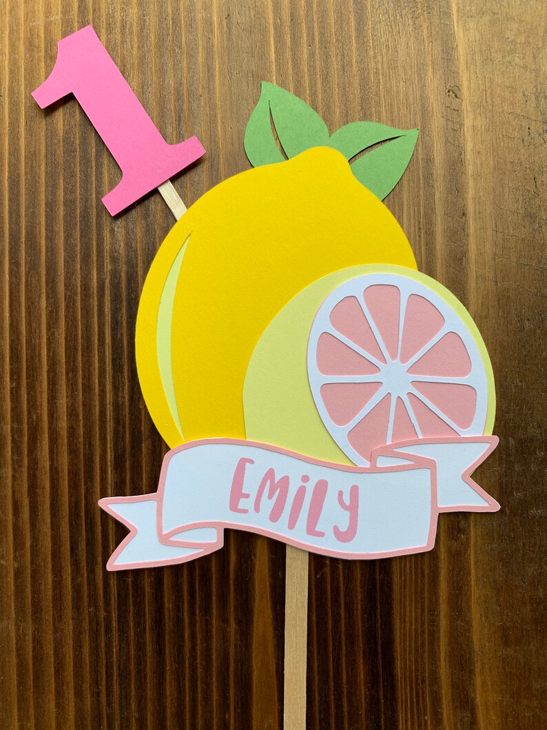 May include: A pink number one cake topper with a yellow lemon and a pink lemon slice. The topper also has a white banner with the name "Emily" written in pink.