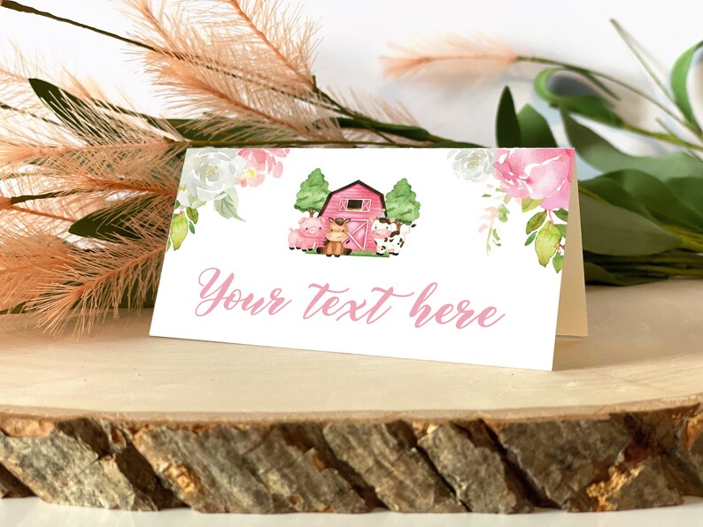 May include: A white folded card featuring a watercolour illustration of a red barn, a pig, a horse, and a cow, accented by pink and white floral arrangements. The text "Your text here" is printed in a pink cursive script on the front of the card.
