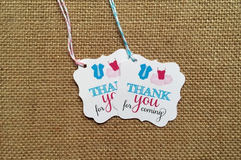 Pode incluir: Duas etiquetas de presente brancas com fita azul e rosa. As etiquetas apresentam um gr&aacute;fico de bailarina e o texto "Obrigado por vir".
