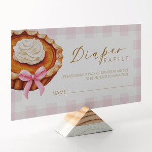 Könnte beinhalten: Eine Babyparty-Windel-Tombola-Karte mit einer Aquarell-Kürbiskuchen-Illustration und einer rosa Schleife. Die Karte trägt den Text "Diaper Raffle" und Anweisungen für die Tombola. Die Karte wird auf einem geometrischen Ständer aus Holz und Weiß angezeigt.