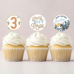 Könnte beinhalten: Drei Cupcakes mit weißem Zuckerguss und dekorativen Tortenauflegern. Einer zeigt die Zahl "3" mit Kakteen, ein anderer "Young Wild & Three" und der letzte "Happy Birthday Bradley" mit einer Cowboy-Illustration.