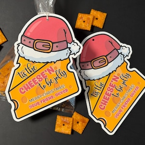 May include: Two gift tags shaped like cheese slices, each adorned with a Santa hat and the text "Tis the CHEESE'N to be jolly." The tags have a festive design with a gold buckle and the message "Merry Christmas! Your friend, Oliver."