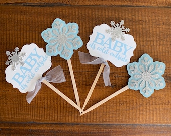 May include: Four blue and silver snowflake themed cupcake toppers with the text "Baby it's cold outside".
